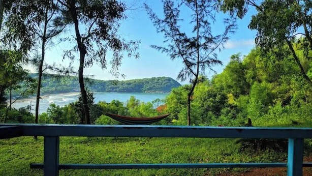 Un hamac est suspendu à une balustrade, offrant une vue sur un paysage verdoyant et luxuriant, avec un plan d’eau et des collines à l’horizon.