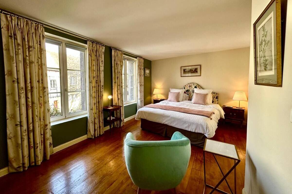 Ein Schlafzimmer mit einem Bett, einem grünen Sessel, einem kleinen Tisch und einem Fenster mit Vorhängen.