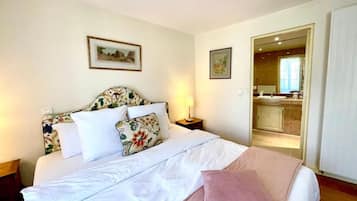 Een net opgemaakt bed met bloemen- en effen kussens, een nachtkastje met een lamp, en uitzicht op een badkamer met een wastafel en spiegel.