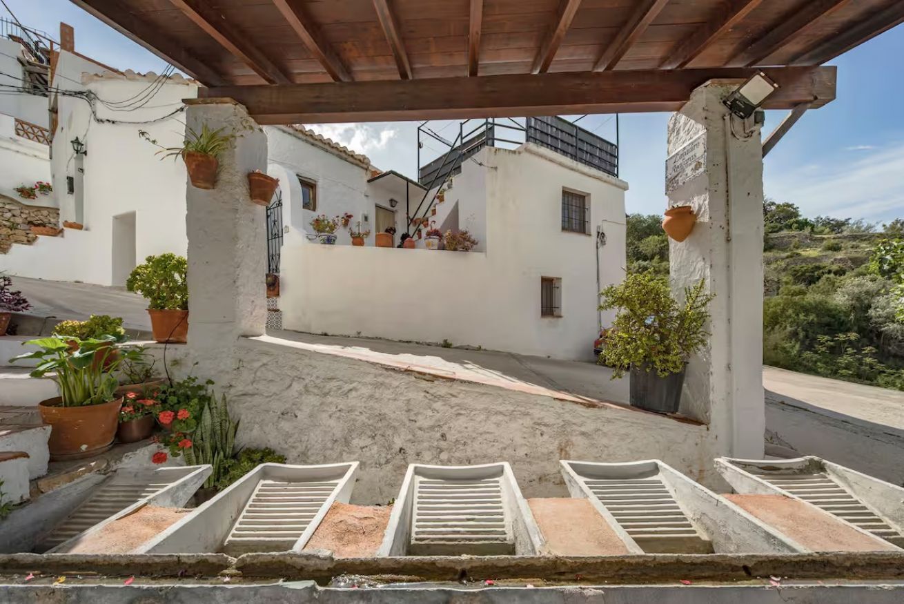 A covered outdoor area with a concrete floor, potted plants, and a staircase leading to a white building.