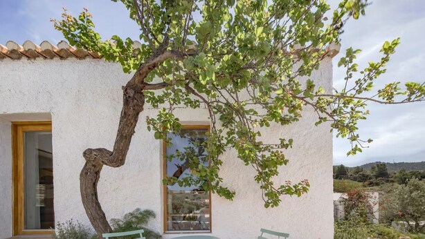A rustic building with a white stucco exterior, a wooden door, and a window. There is a small round table with two chairs outside. - El Castillete – Charm with Sea Views (La Garnatilla)
