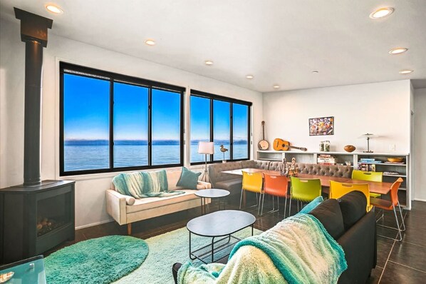 A modern living room with a large window overlooking the sea, a fireplace, a dining area with colorful chairs, and a cozy seating area.