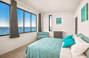 A bedroom with a large bed, a view of the sea, a chair, a dresser, and a painting on the wall.