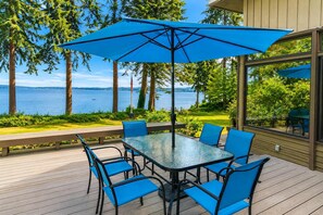 A deck with a glass-top table and blue chairs, overlooking a lake with trees and a distant view of a city skyline. - Poulsbo's Waterfront Lodge · Puzzle Point (Poulsbo)