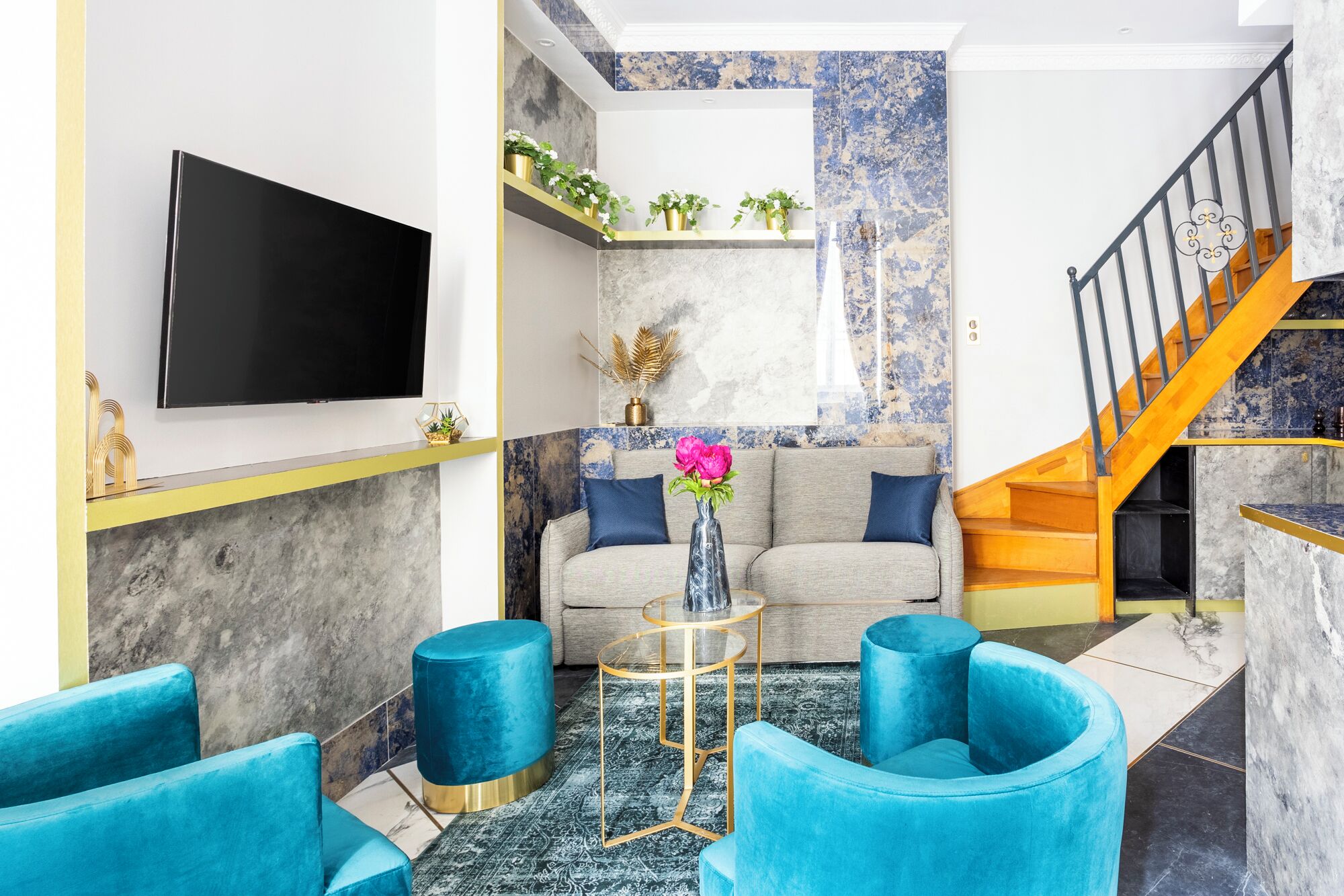 A living room with a sofa, two blue armchairs, a coffee table, and a TV. There is a staircase with a wooden railing and a blue and white patterned wall.
