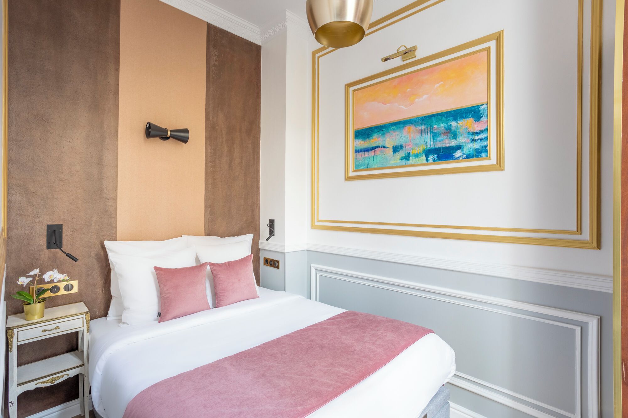 A hotel room with a bed, a nightstand, a wall-mounted lamp, and a painting.