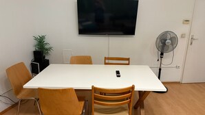 Una sala de conferencias con una mesa blanca grande, seis sillas de madera, un televisor montado, un ventilador de pie y una planta en maceta.