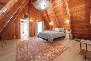 A cozy attic bedroom with a sloped ceiling, wooden walls, a bed with a gray blanket, a bedside table, and a patterned rug.