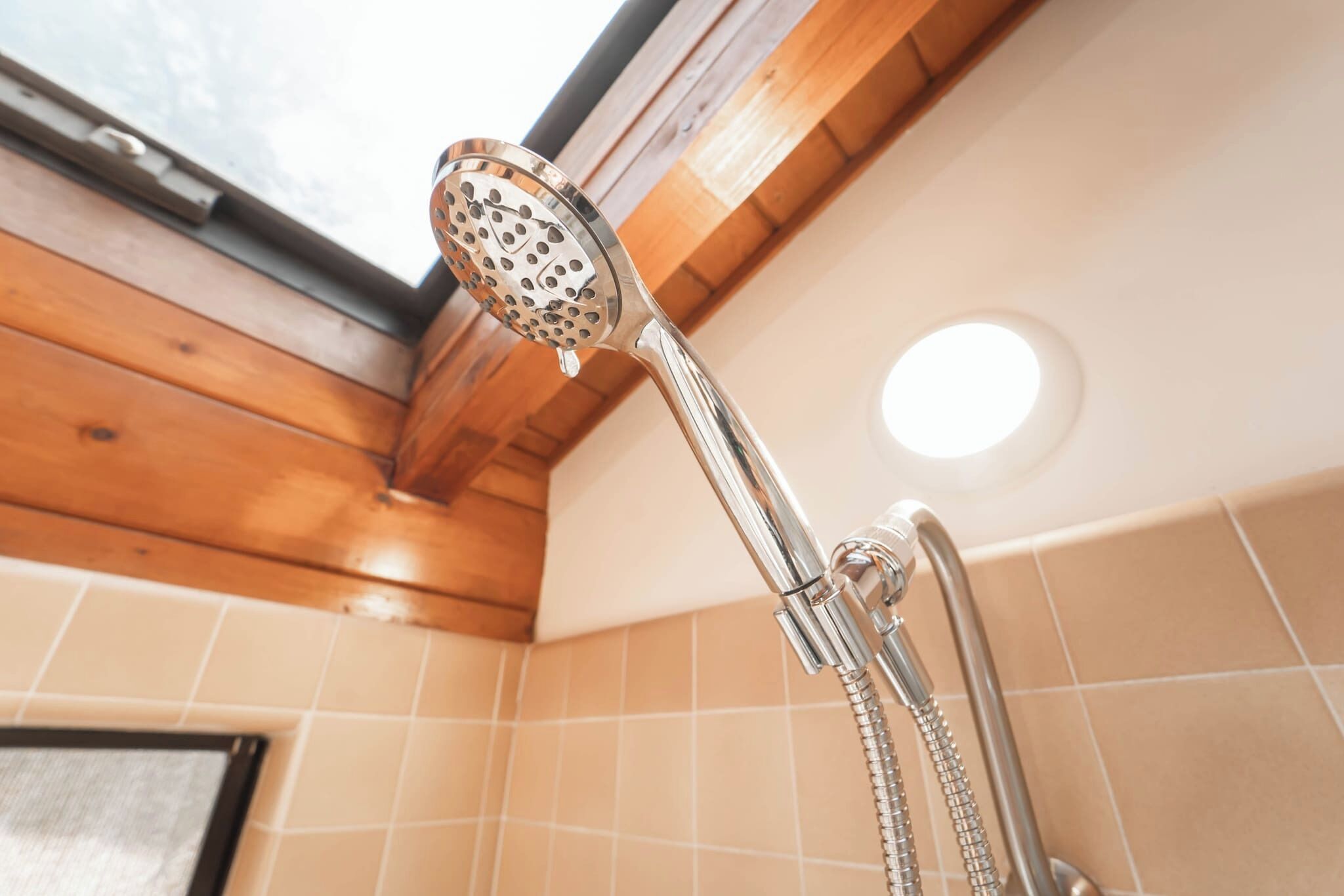 Un cabezal de ducha con un accesorio tipo lluvia en un cuarto con vigas de madera y baldosas color crema.
