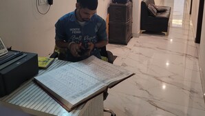 A person seated at a desk with a laptop, papers, and a mobile phone. The room has a tiled floor, a framed picture on the wall, and a bench in the background. - Roomshala Hotel Scorpio (New Delhi)
