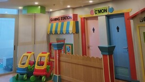 Espaço infantil com área de jogos, um aviso 'C'MON' e uma porta azul com símbolos de banheiro masculino e feminino.