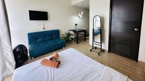Quarto de hotel com cama, sofá azul, escrivaninha com cadeira, TV na parede, espelho com rodízios e porta escura.