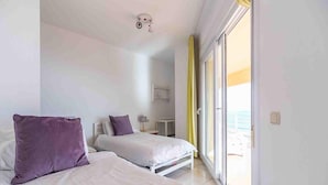 A hotel room with two beds, a bedside table, a lamp, and a view of the sea through a sliding door. - Mediterranean Beach front Apartment: Large terrace, Air Conditioning, Wi-fi, Pool. (Fuengirola)