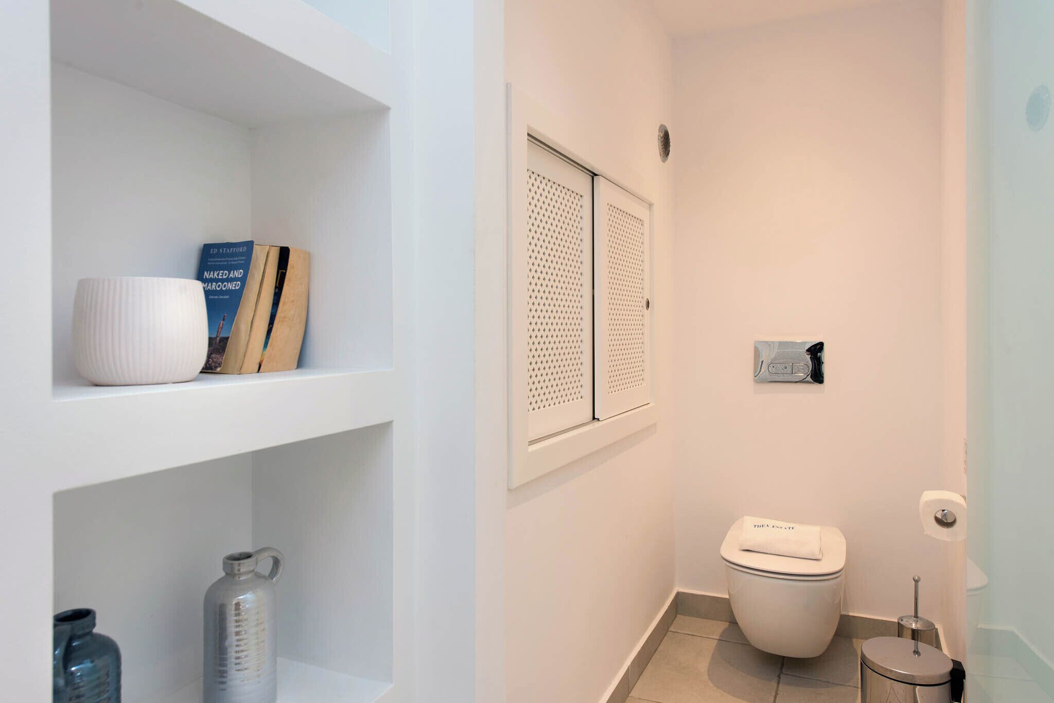 Un baño pequeño y limpio con inodoro, una repisa con libros y una botella de agua, y un dispensador de papel higiénico en la pared.