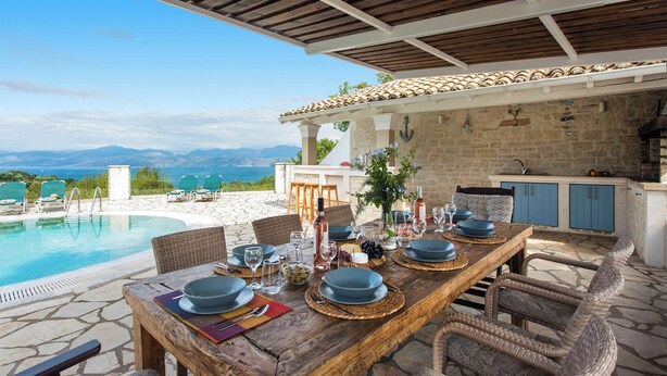 A wooden dining table set for six with blue plates, surrounded by wicker chairs, overlooking a pool and mountains. - Thea Estate (Corfu)