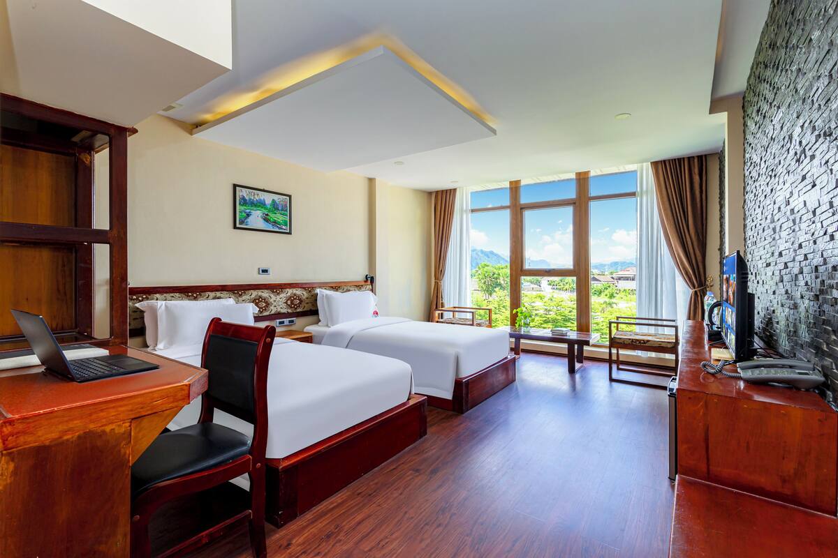 A hotel room with two beds, a desk with a laptop, a wooden chair, a desk lamp, a telephone, and a large window with a view of greenery.