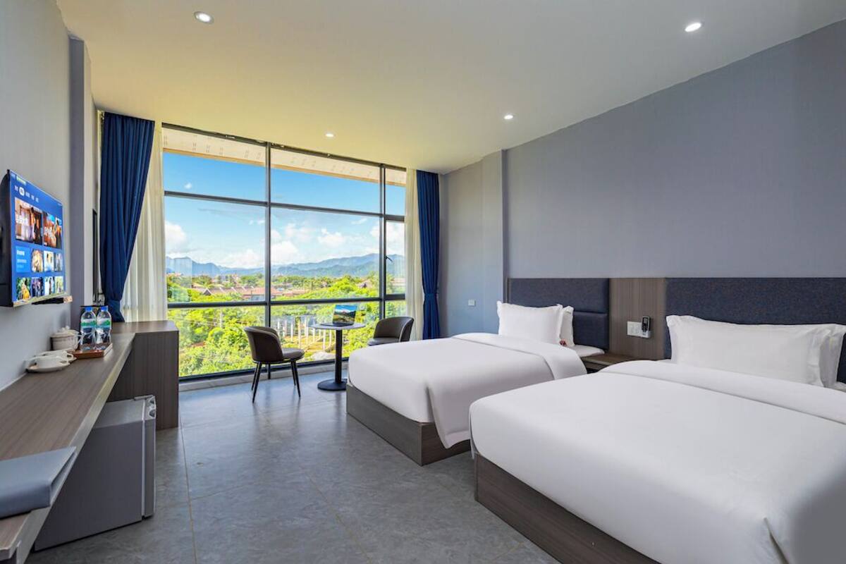 A hotel room with two beds, a desk with a computer, a small table, and a large window offering a view of the outdoors.