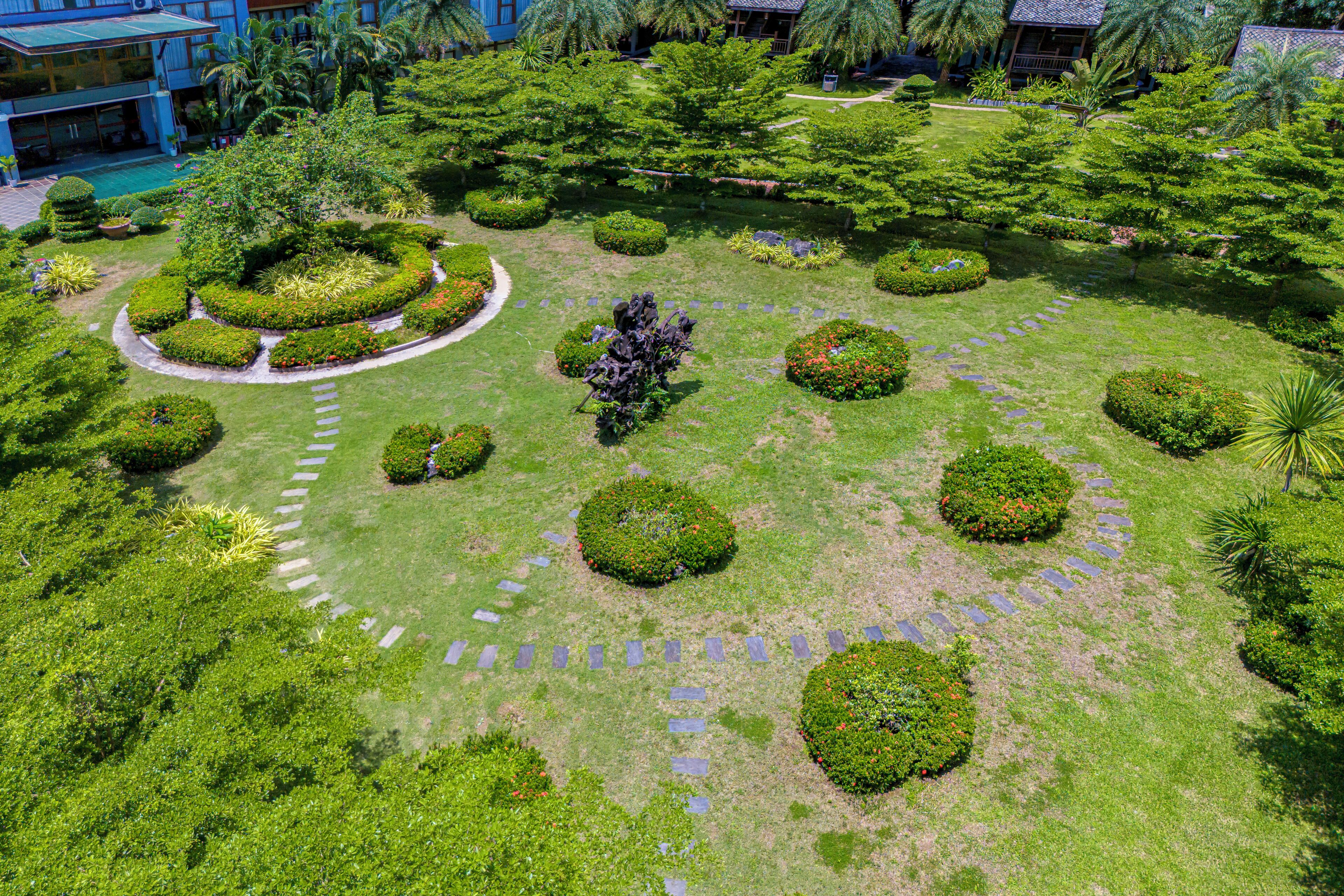 An aerial view of a well-maintained garden with circular and oval flower beds, a central statue, and a paved walkway.