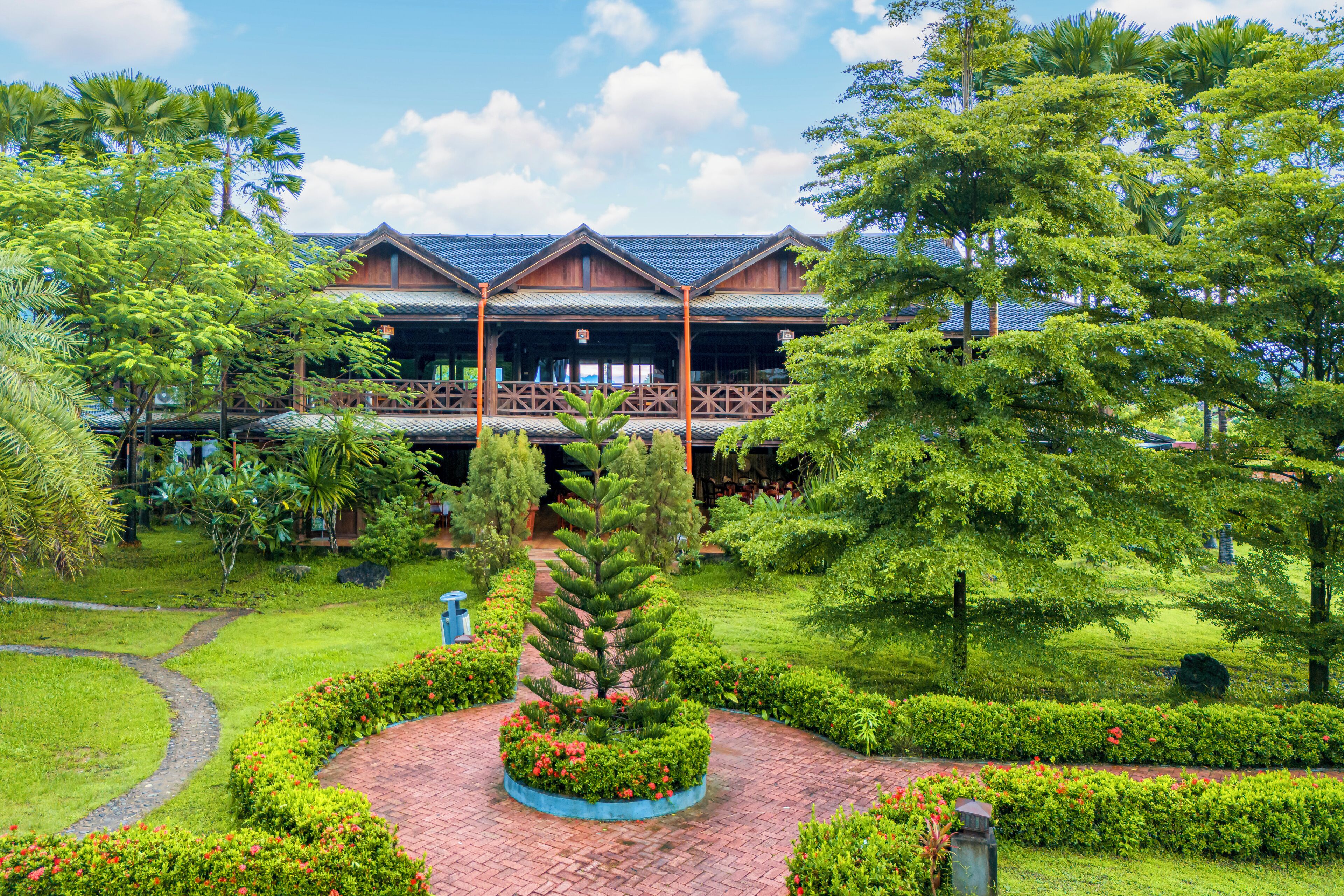 Un hôtel à l’architecture traditionnelle, avec une structure en bois, entouré d’une végétation luxuriante et d’un jardin bien entretenu.