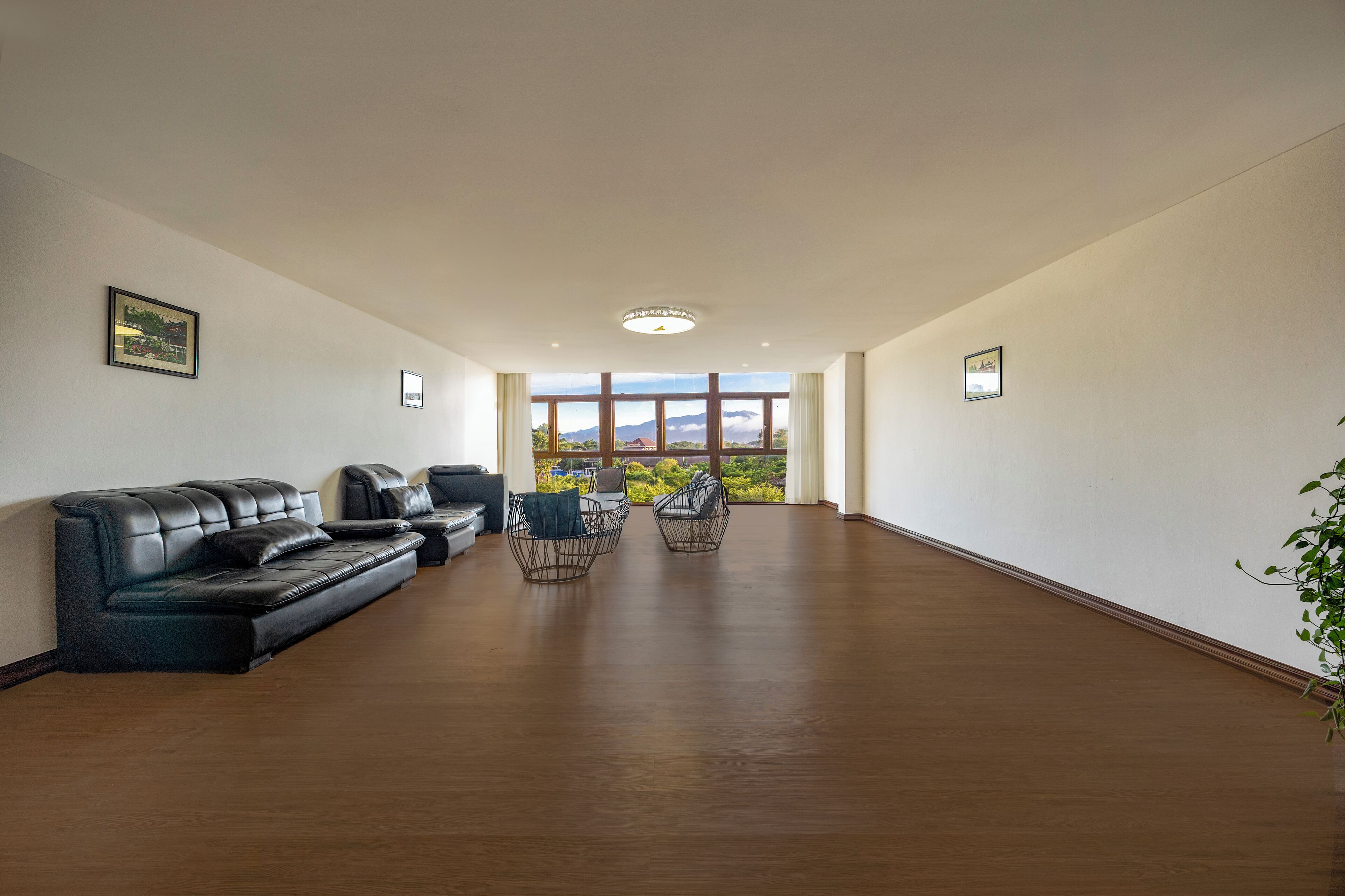 A spacious room with a wooden floor, a black leather sofa, two wireframe chairs, and a view through large windows.