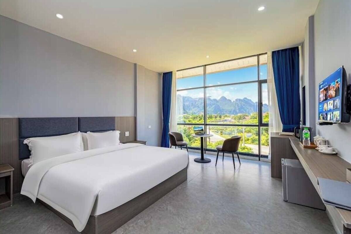 A modern hotel room with a large bed, a small desk, a chair, and a view of mountains through large windows.