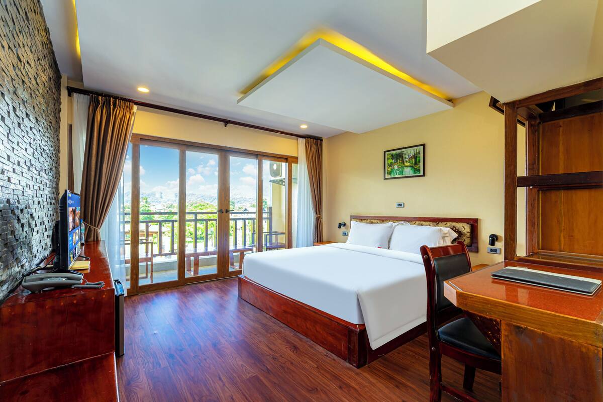 A hotel room with a large bed, wooden furniture, a desk, and a balcony with a view.