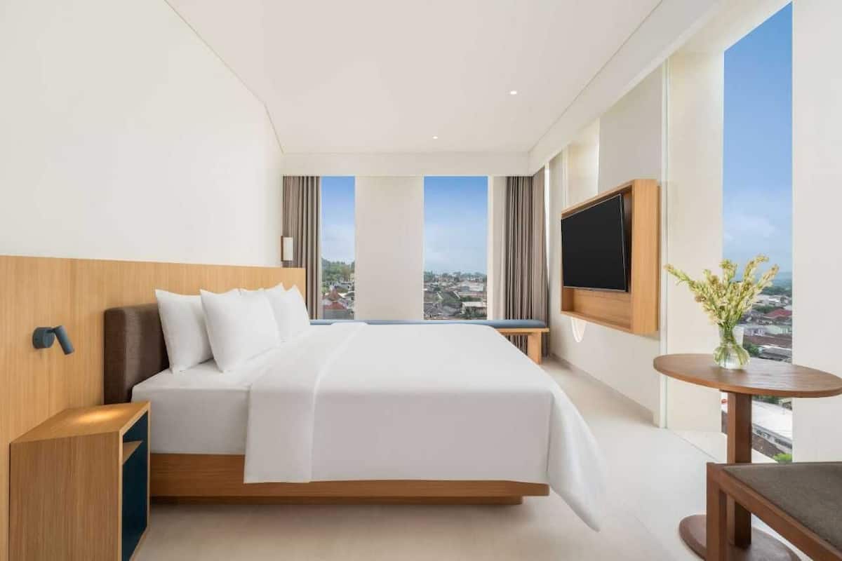 Habitación de hotel moderna con una cama grande, televisor montado en la pared, una mesita con un jarrón y vistas a la ciudad a través de amplios ventanales.