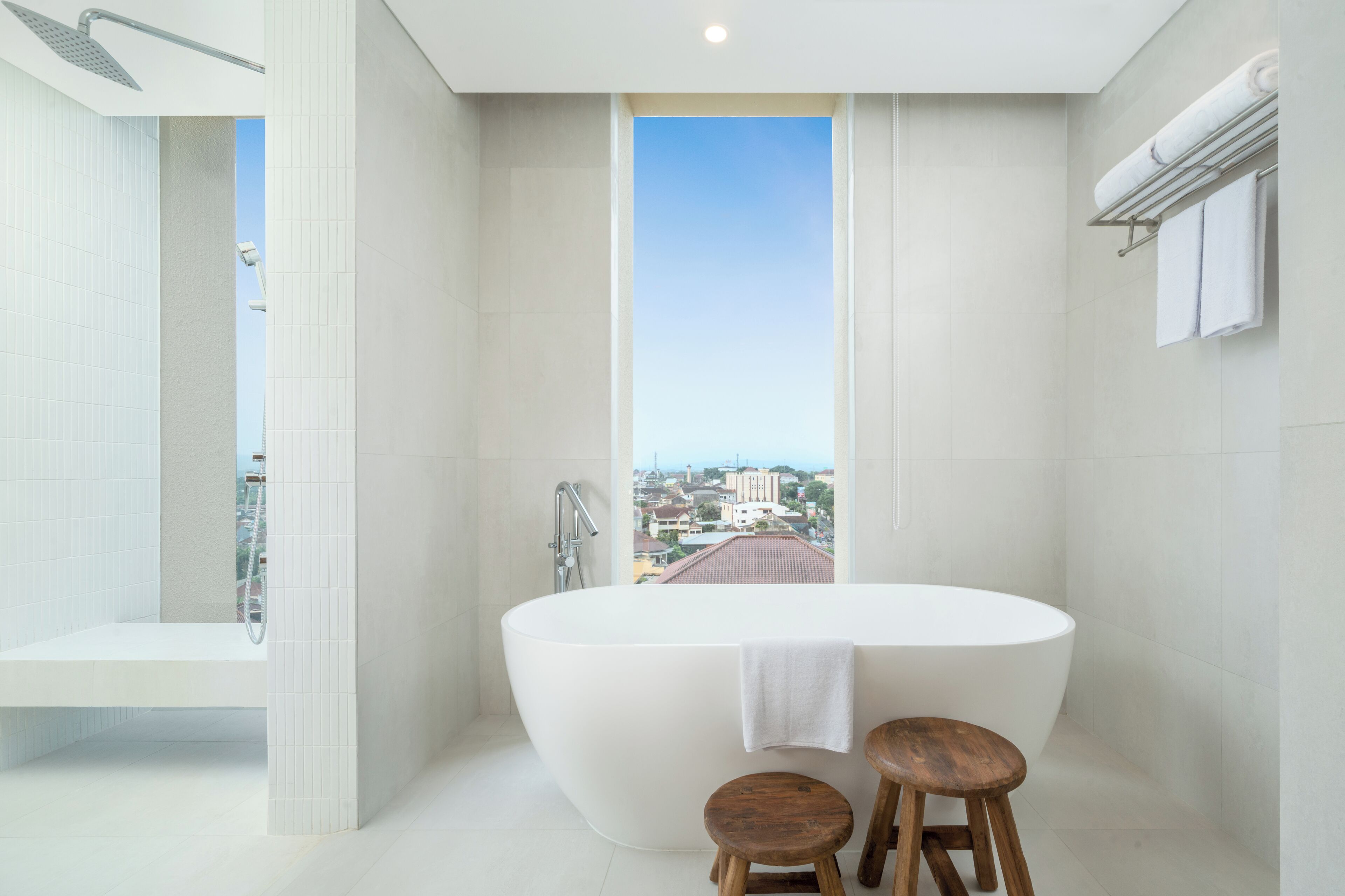 Ein modernes Badezimmer mit einer freistehenden Badewanne, zwei hölzernen Hockern, einem Handtuchhalter und Blick auf die Stadt durch ein großes Fenster.
