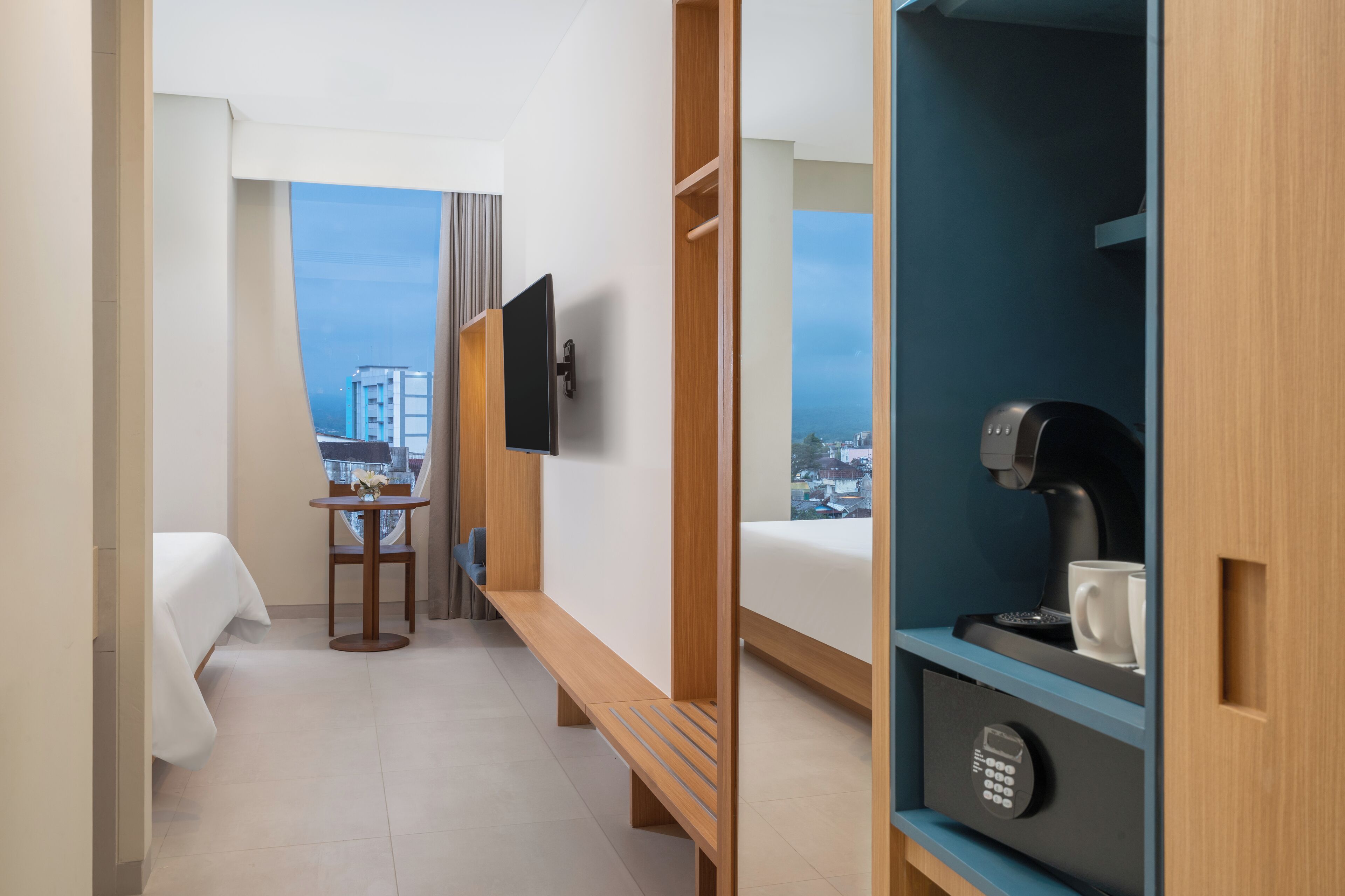 Habitación de hotel con cama, una mesita con silla, televisor montado en la pared, cafetera y vistas a la ciudad.