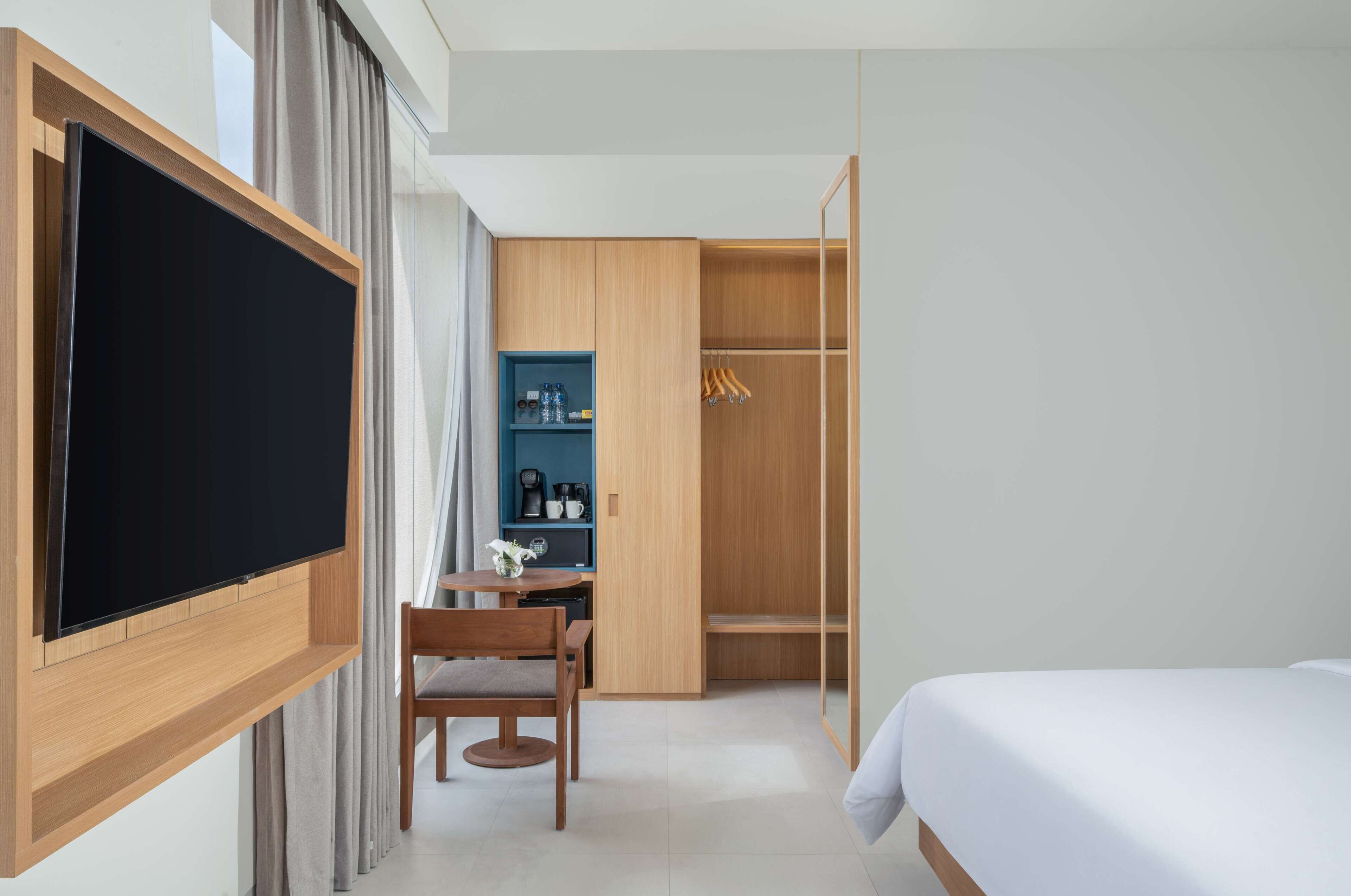 A hotel room with a bed, a TV mounted on the wall, a desk with a chair, a wardrobe, and a small table with a vase.
