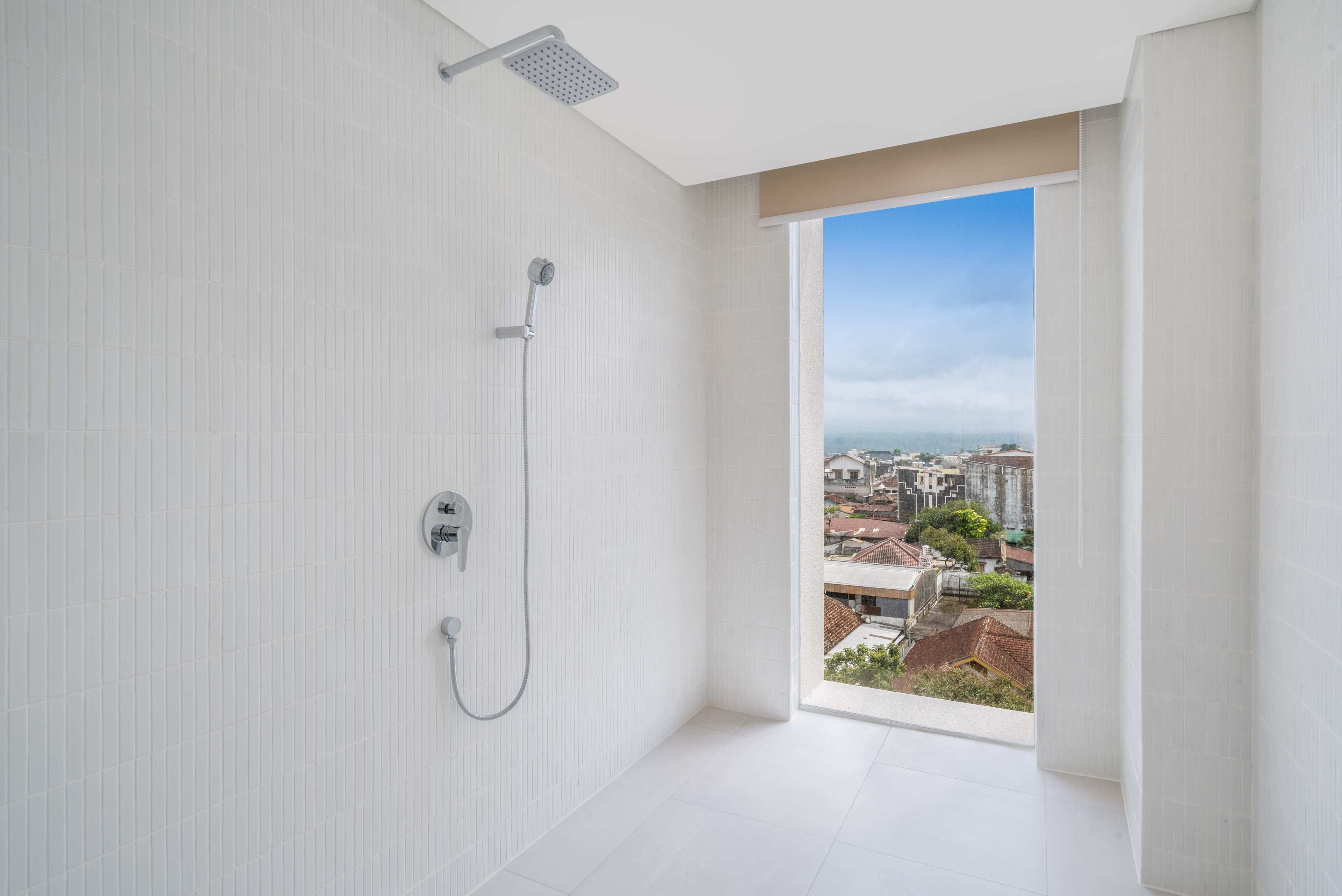 A modern bathroom with a shower, a view of the city through a large window, and a clean, white tiled floor.
