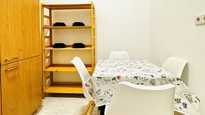 Een kleine kamer met een houten kast, een witte tafel met een bloemig tafelkleed, en witte stoelen.