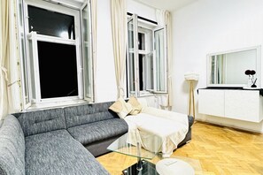 A living room with a grey sofa, a glass coffee table, a white cabinet, and a large window with curtains.