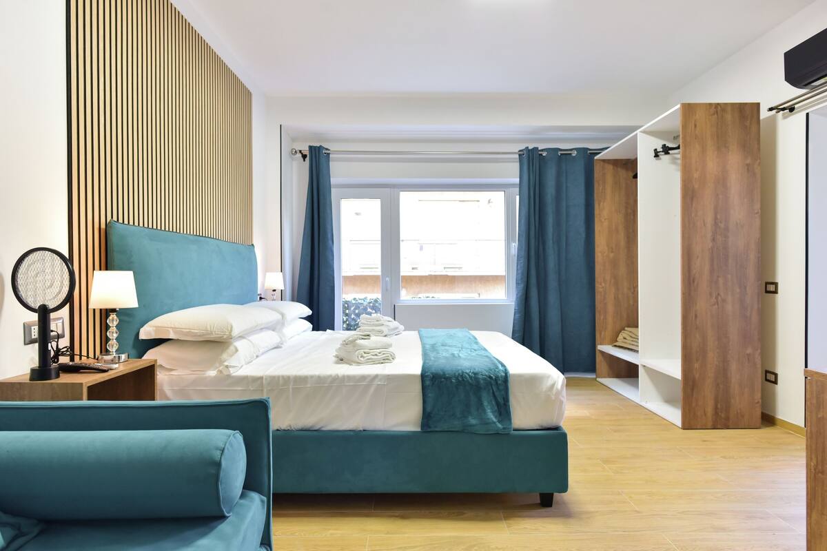 A modern hotel room with a large bed, a bedside table with a lamp, a chair, and a view of the outdoors through a window.