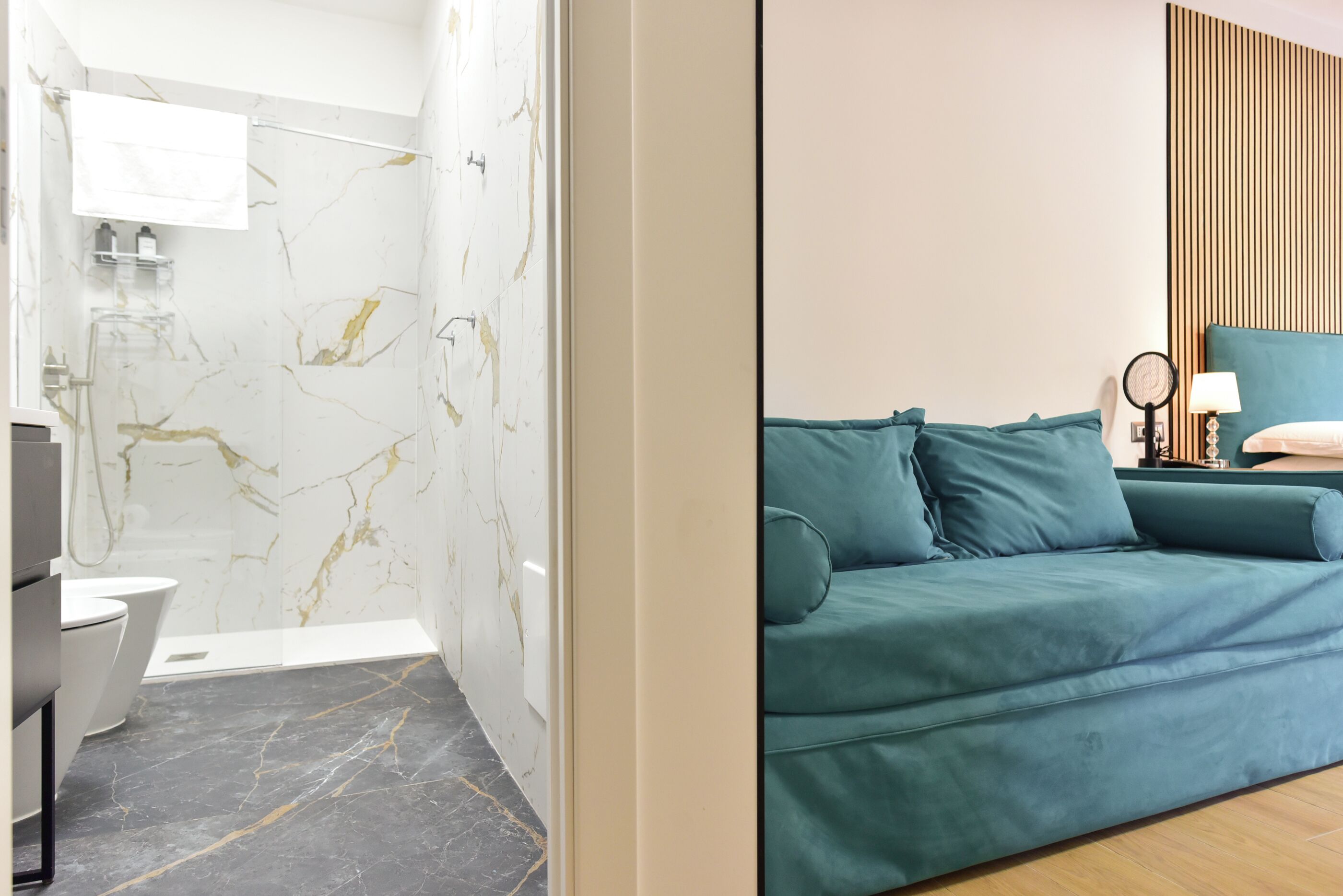 A modern bathroom with a marble wall, a toilet, and a shower. There is a teal sofa with a lamp and a small table in the living area.