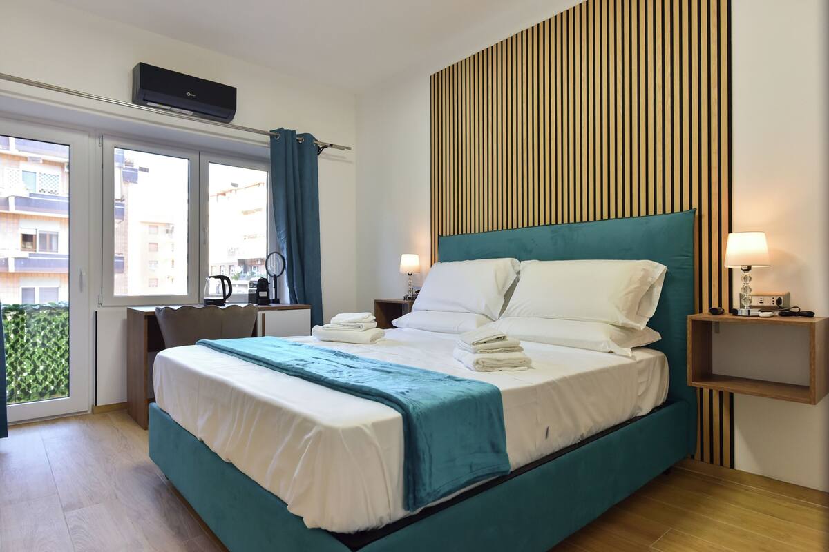 A modern hotel room with a large bed, a desk, and a view of the city through the windows.