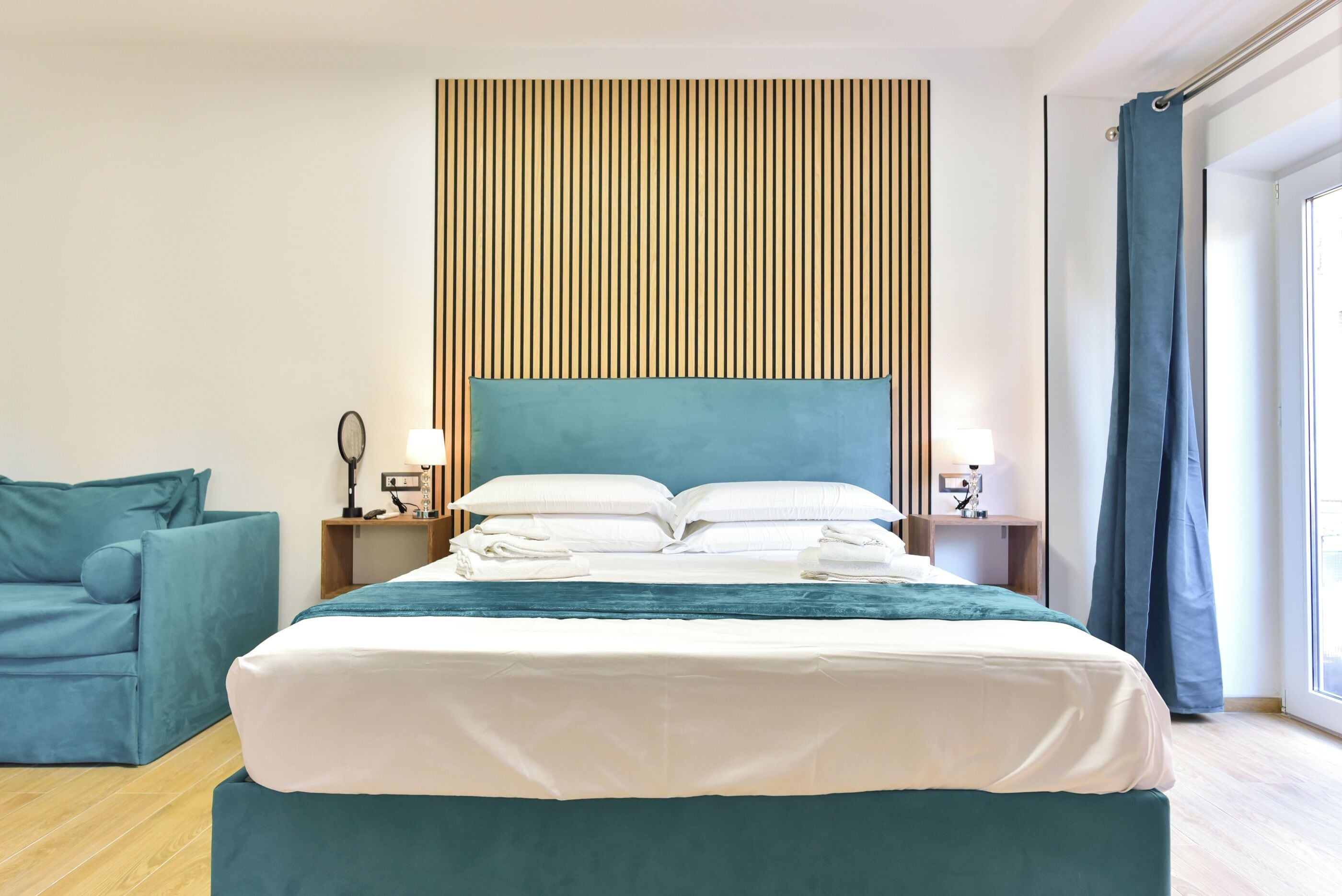 A modern hotel room with a large bed, a blue headboard, a bedside table with lamps, and a view of the outdoors.
