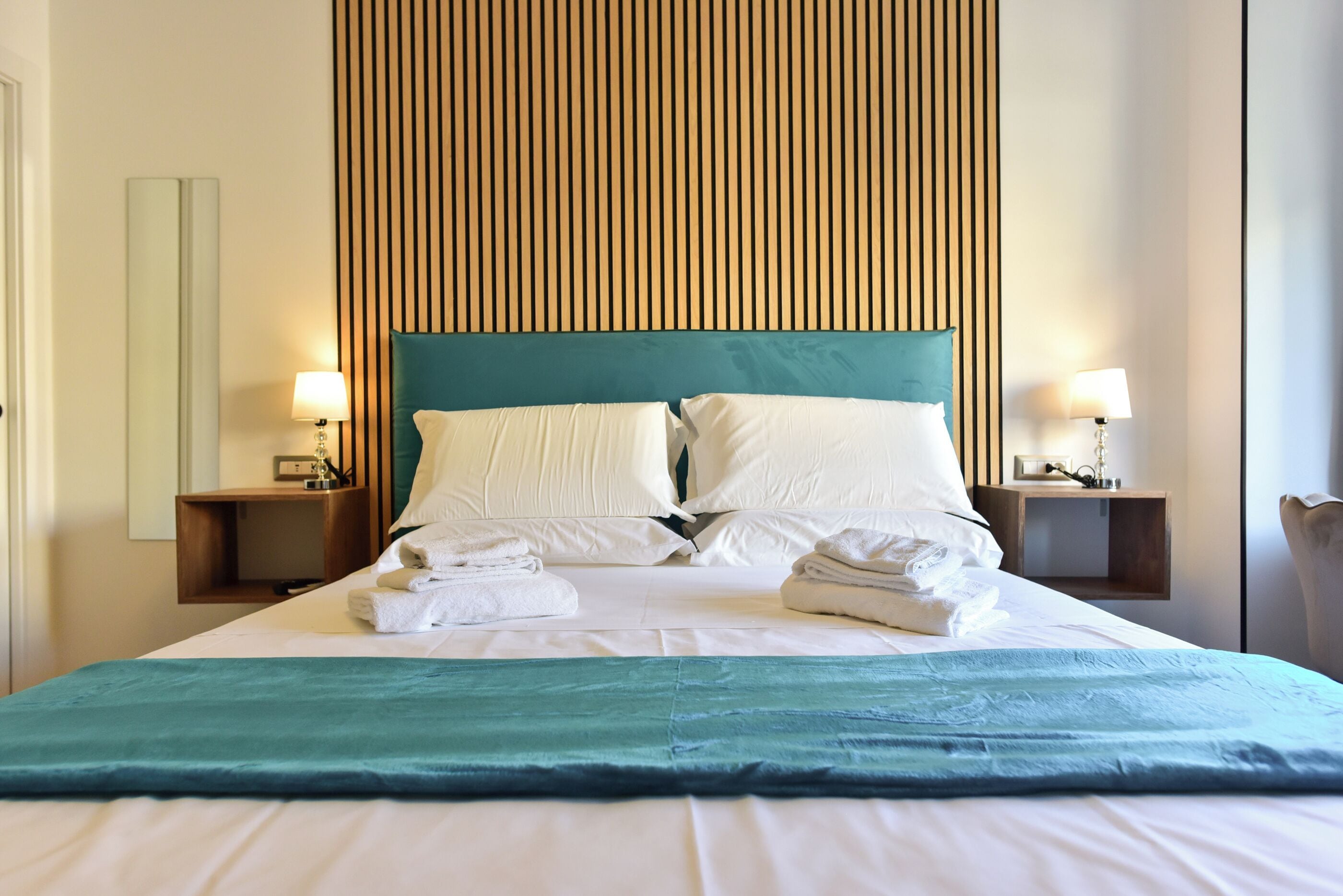 A neatly made bed with white linens and a teal throw blanket, flanked by two bedside tables with lamps, in a room with a wooden slat headboard.