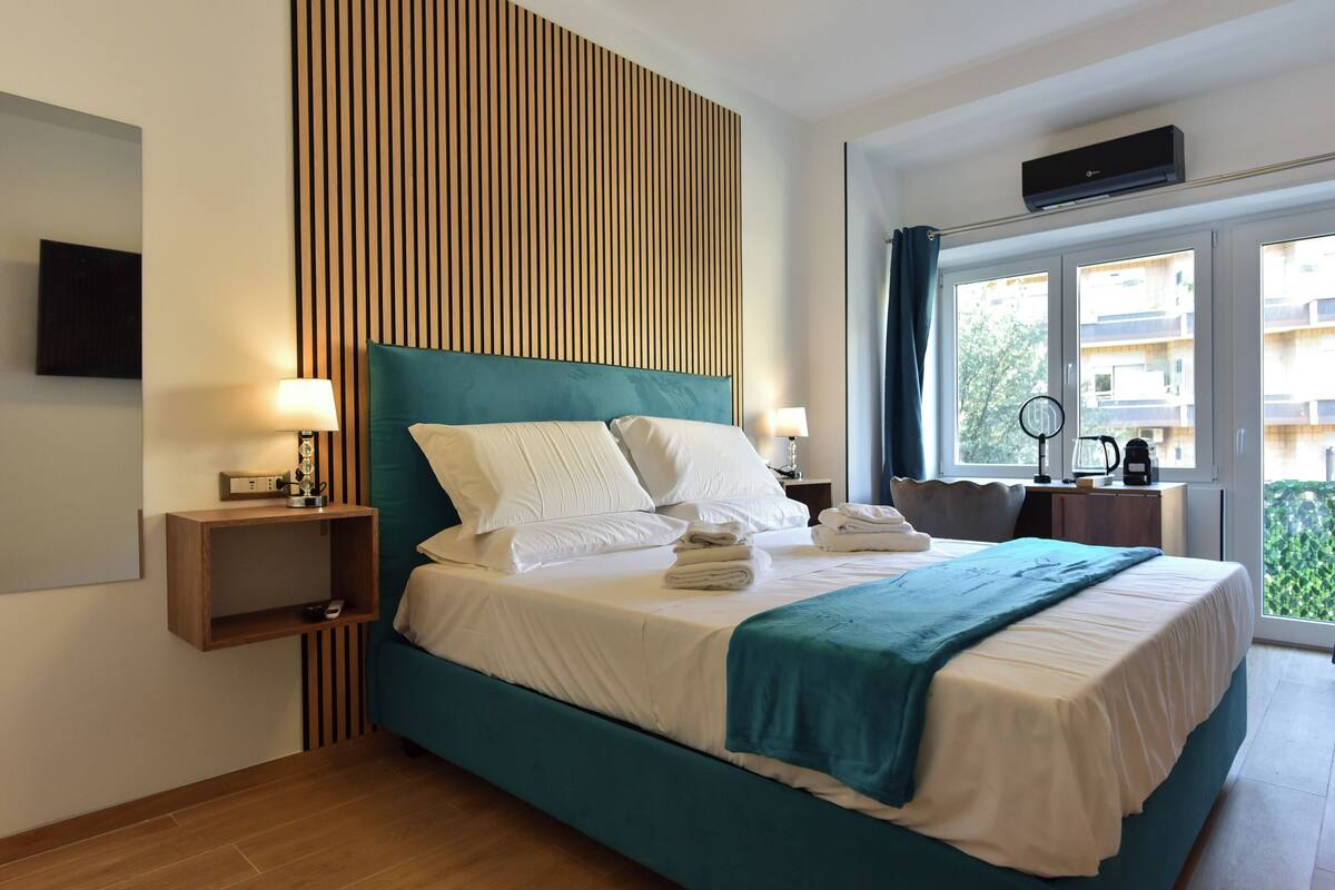 A hotel room with a large bed, a wooden slat wall, a TV, a bedside table with a lamp, and a window with a view of buildings.