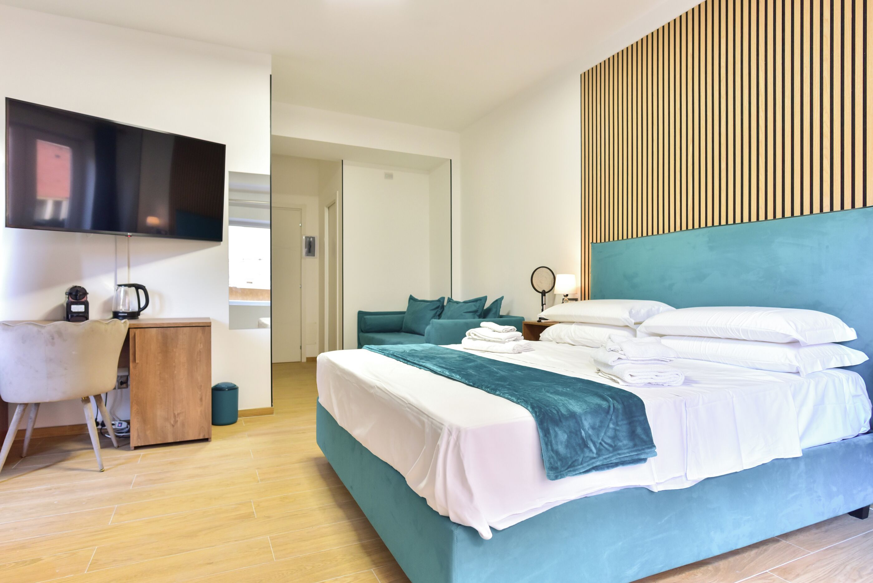A modern hotel room with a large bed, a desk with a kettle, and a flat-screen TV mounted on the wall.