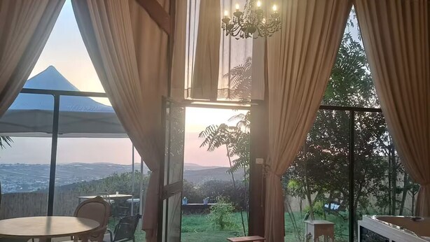 Il s’agit d’une pièce spacieuse avec une grande fenêtre donnant sur une montagne et un coucher de soleil. On y trouve une table à manger avec des chaises, un canapé et un téléviseur fixé au mur.
