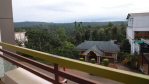 A view from a balcony overlooking a residential area with a house, greenery, and a distant body of water. - Hotel Mountain Hues Thekkady (Udumbanchola)