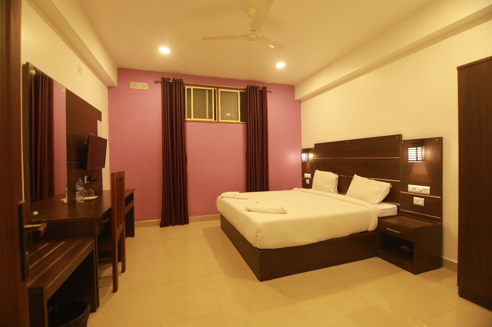 A hotel room with a bed, bedside table, desk, chair, curtains, and a ceiling fan.