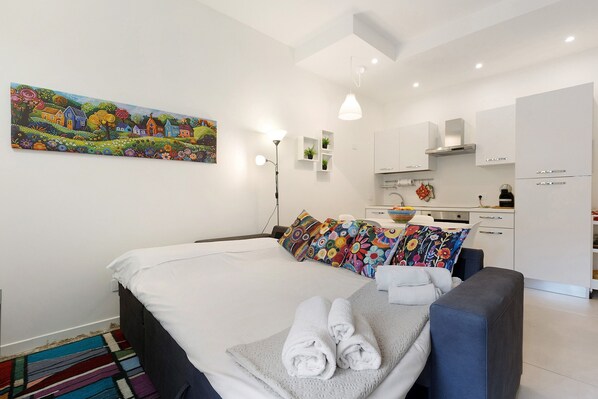 A modern hotel room with a bed, a sofa, a colorful painting, and a kitchenette.