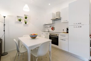A modern kitchen with white cabinetry, a dining table with chairs, a bowl of fruit, and a pendant light.