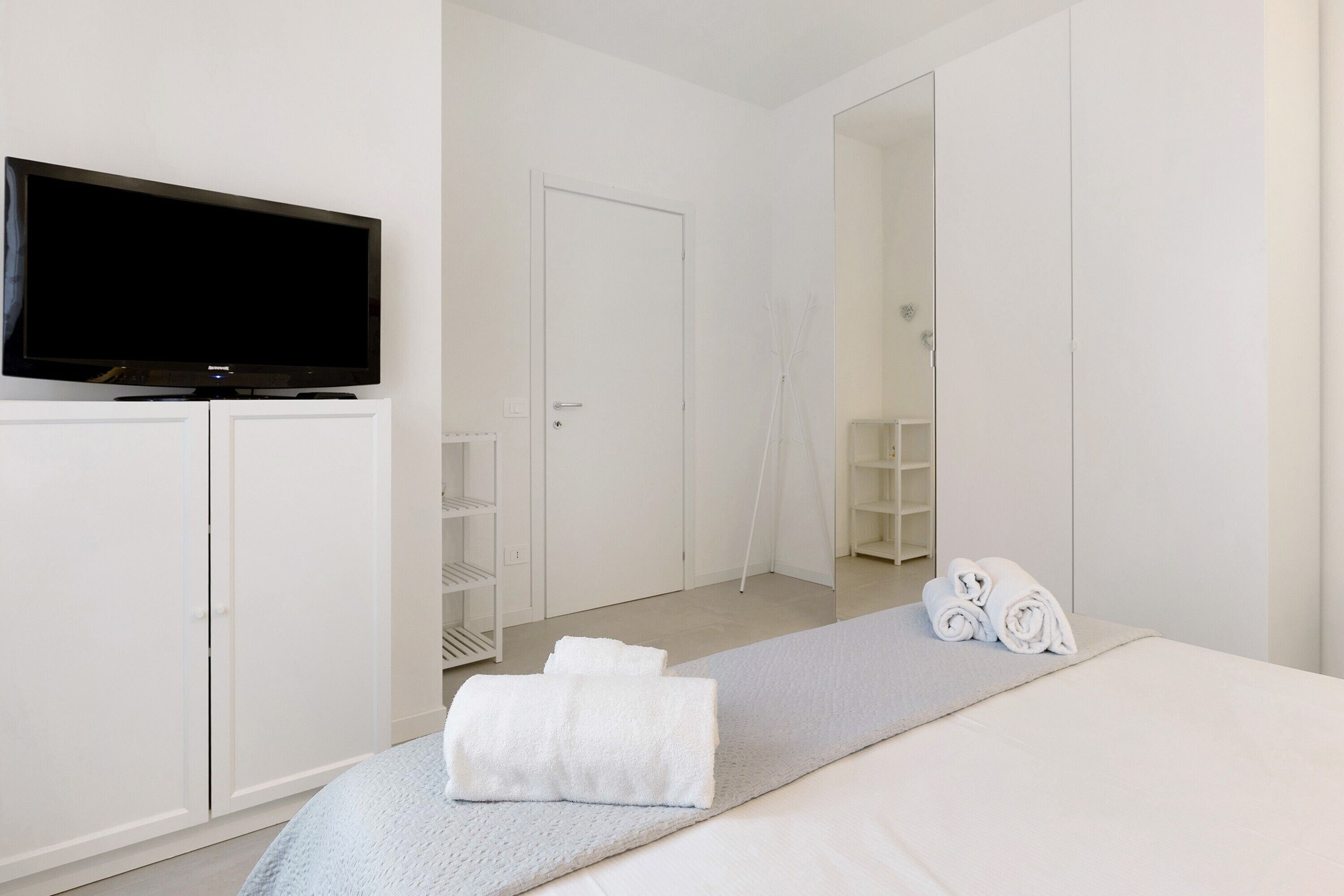 A hotel room with a bed, a TV on a cabinet, a mirror, and a towel rack.
