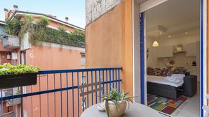 A balcony with a potted plant, a small table, and a view into a room with a bed, a sofa, and a kitchen area.