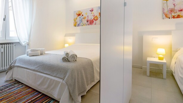 A hotel room with a bed, a bedside table with a lamp, a colorful rug, and a window with curtains.