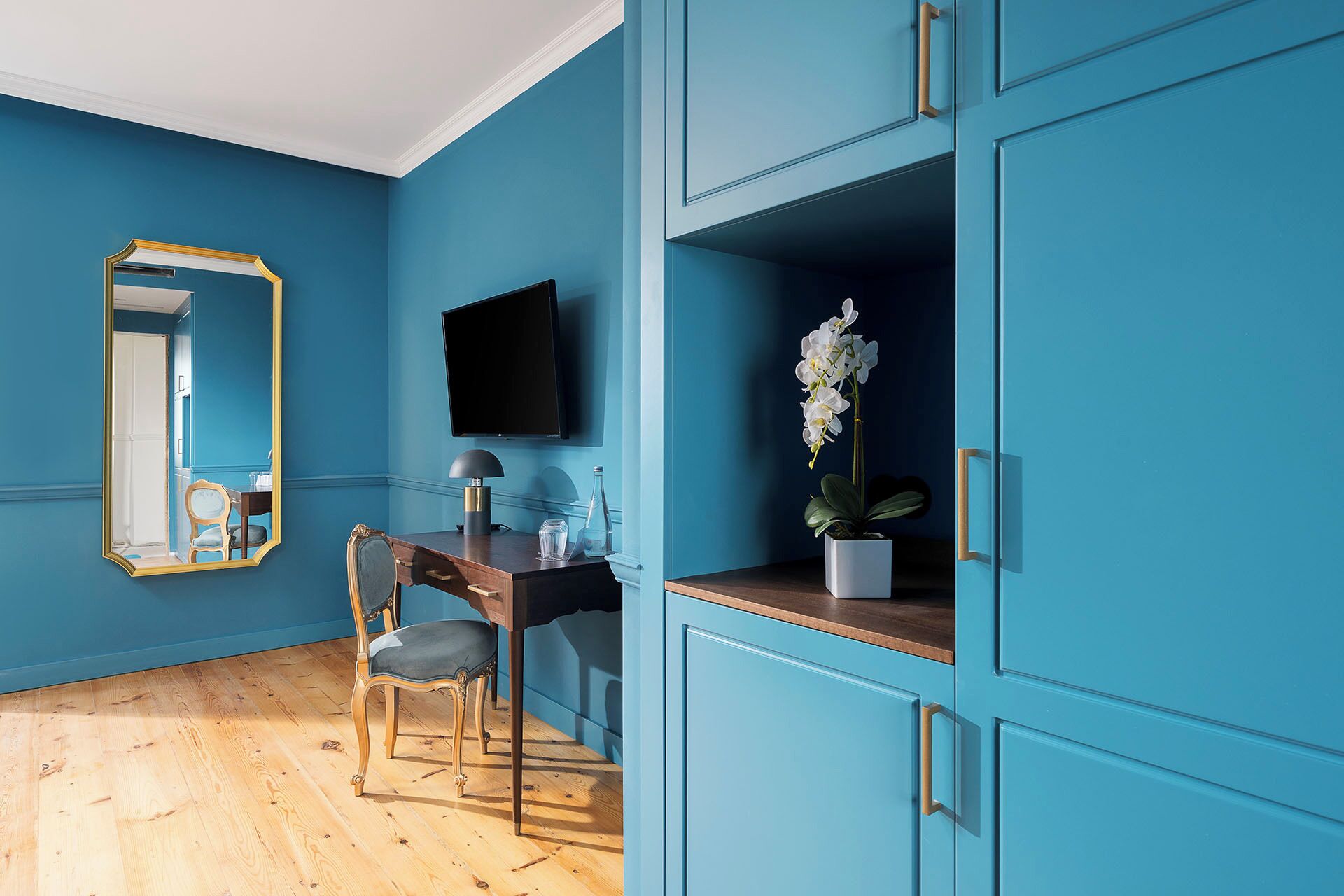 A room with teal walls, a wooden floor, a desk with a chair, a television, and a potted orchid.