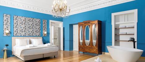 A bedroom with a large bed, a wooden armoire, a freestanding bathtub, and a chandelier.